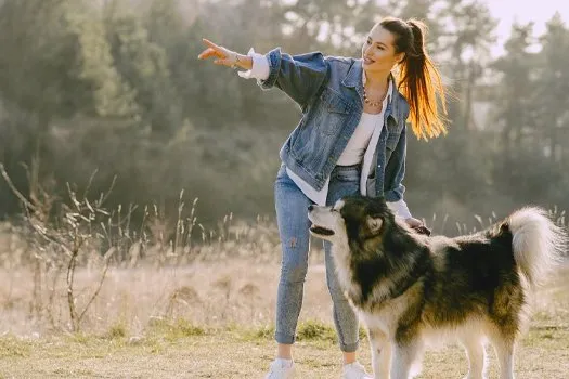 Kot ceketli bir kadının elini yana doğru uzatıp işaret ettiği, yanında duran tüylü siyah-beyaz bir köpeğin fotoğrafı