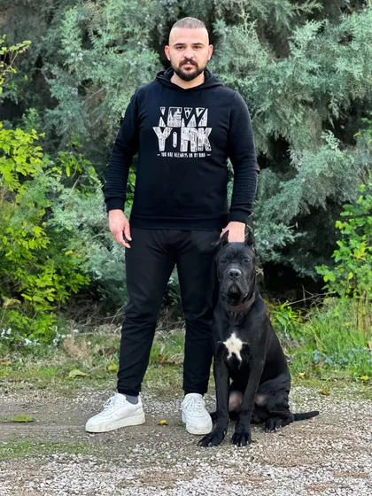 Çam ağaçları önünde ayakta duran bir adamın yanında, göğsünde beyaz lekesi olan siyah bir Cane Corso köpeğinin oturduğu fotoğrafı